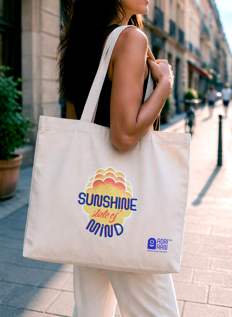 katoenen draagtas met “Sunshine state of mind” print en klein ADRIAAN vzw logo, gedragen over de schouder in een winkelstraat