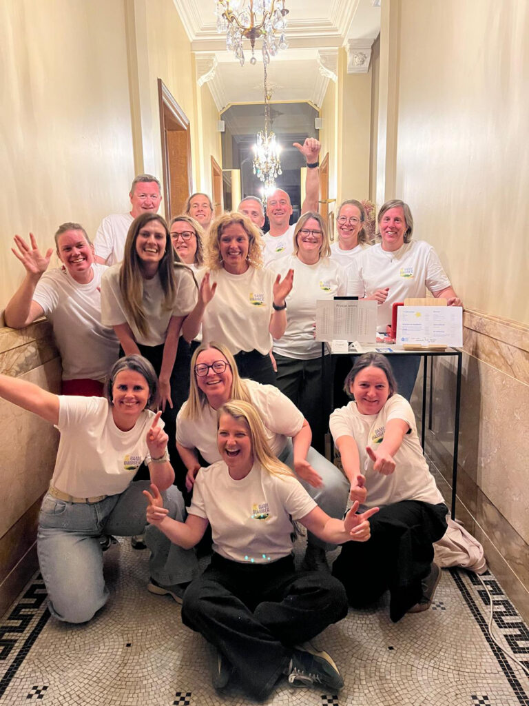 Groep van vijftien vrolijke mensen in witte “The Game Changer club” T-shirts poseert juichend in een statige gang met kroonluchters