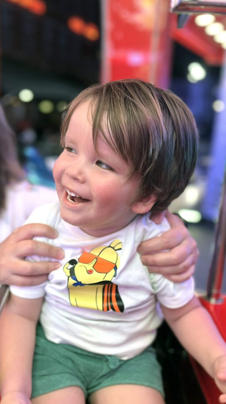 Adriaan lacht en kijkt opzij terwijl iemand hem vasthoudt, in een drukke, kleurrijke kermis