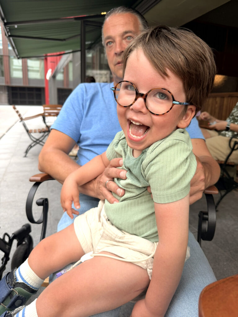 Adriaan met bril lacht uitbundig terwijl een man hem op schoot houdt, op een terras