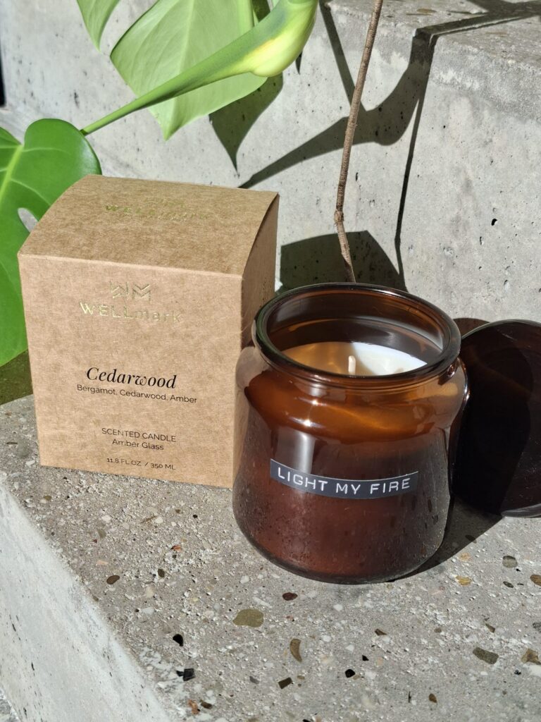 Geopende WELLmark Cedarwood geurkaars in amberglas met losse deksel, naast de kraftkartonnen verpakking